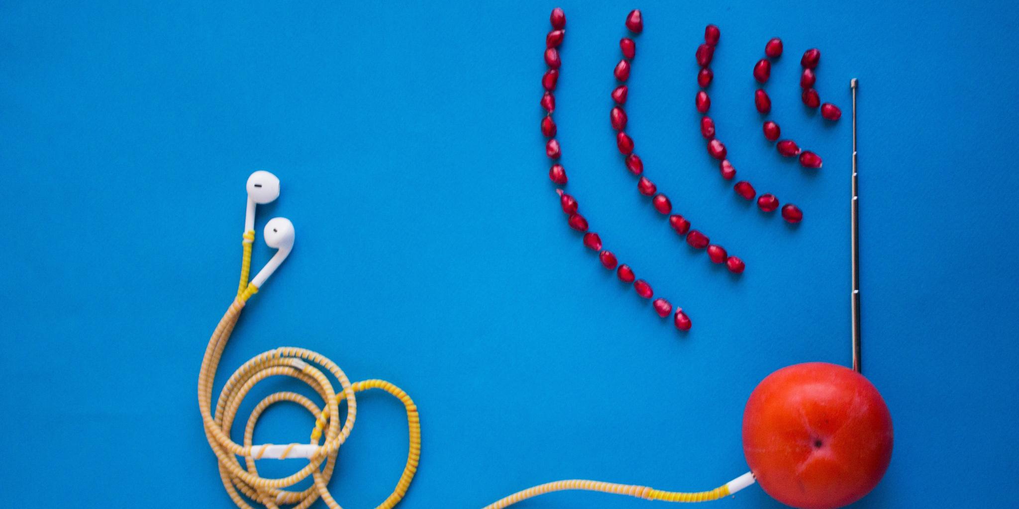 auf einer blauen Fläche liegt eine Mandarine, in der eine Antenne steckt. Vor der Antenne liegen  (in Form von Radiowellen) Granatapfelkerne. Von der Mandarine führt ein Kabel mit Kopfhöhrern, aus Nudeln.