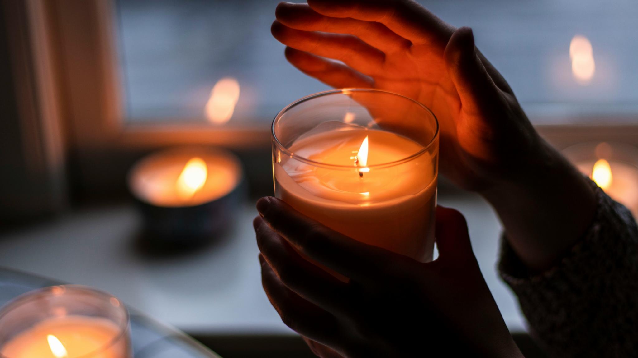 Eine Hand hält eine brennende Kerze in einem Glas und die andere Hand ist schützend über die Flamme gehalten