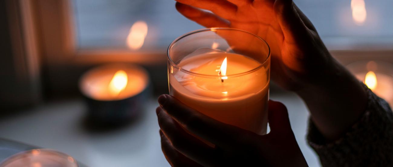 Eine Hand hält eine brennende Kerze in einem Glas und die andere Hand ist schützend über die Flamme gehalten