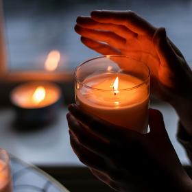 Eine Hand hält eine brennende Kerze in einem Glas und die andere Hand ist schützend über die Flamme gehalten