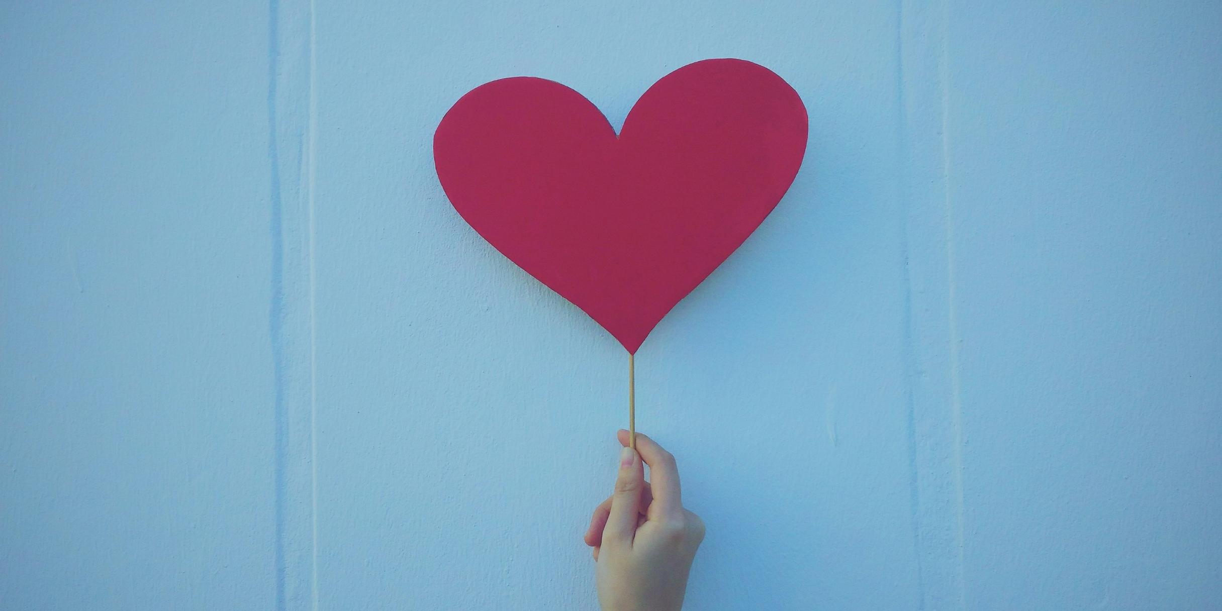Eine Hand hält ein Herz an einem Stiel vor einer Wand nach oben