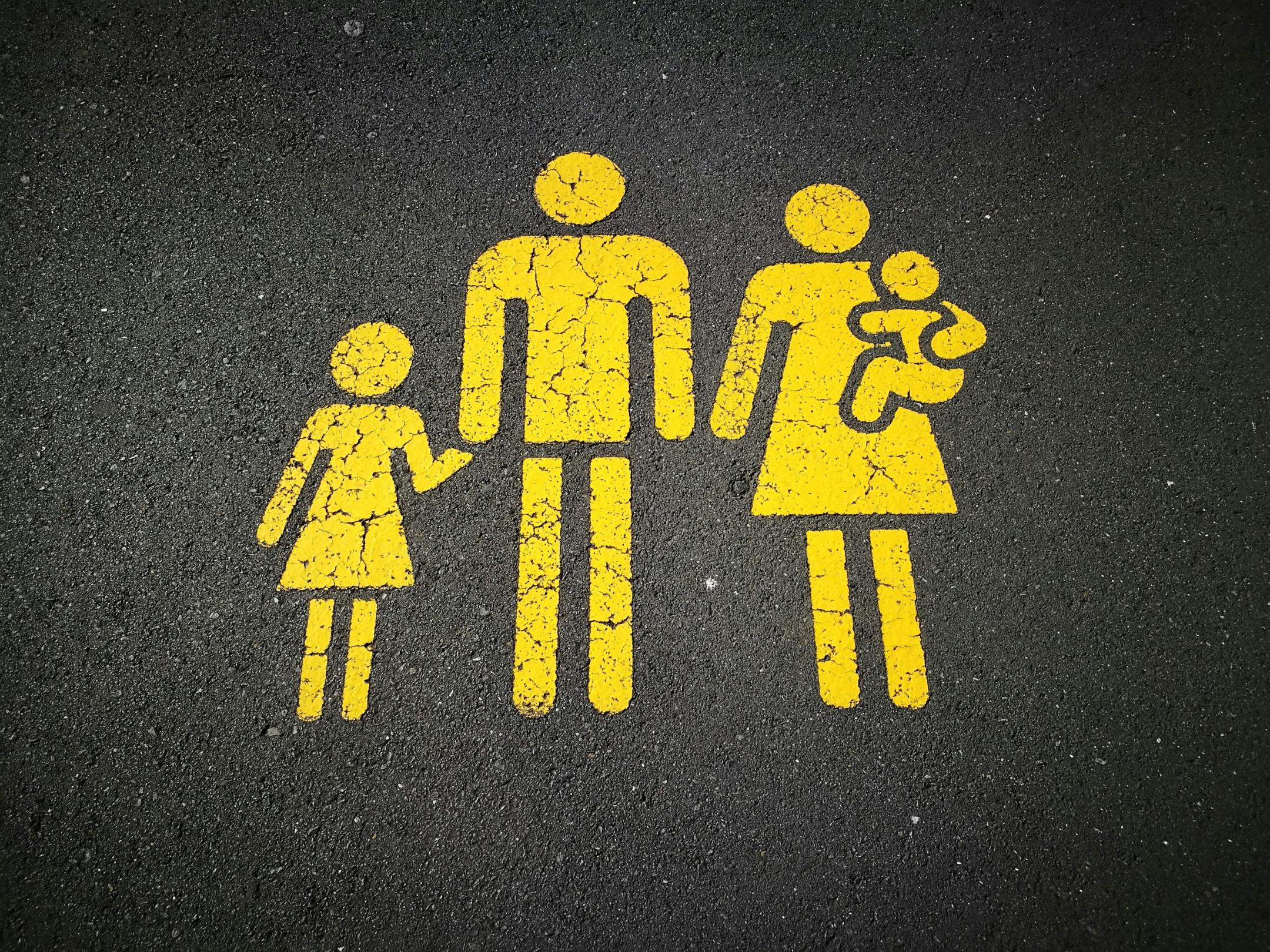 Auf einer Straße sind gelbe Strichmännchen aufgezeichnet. Es ist eine Familie von denen der Mann ein Kind an der Hand hält und die Frau ein Kind auf dem Arm