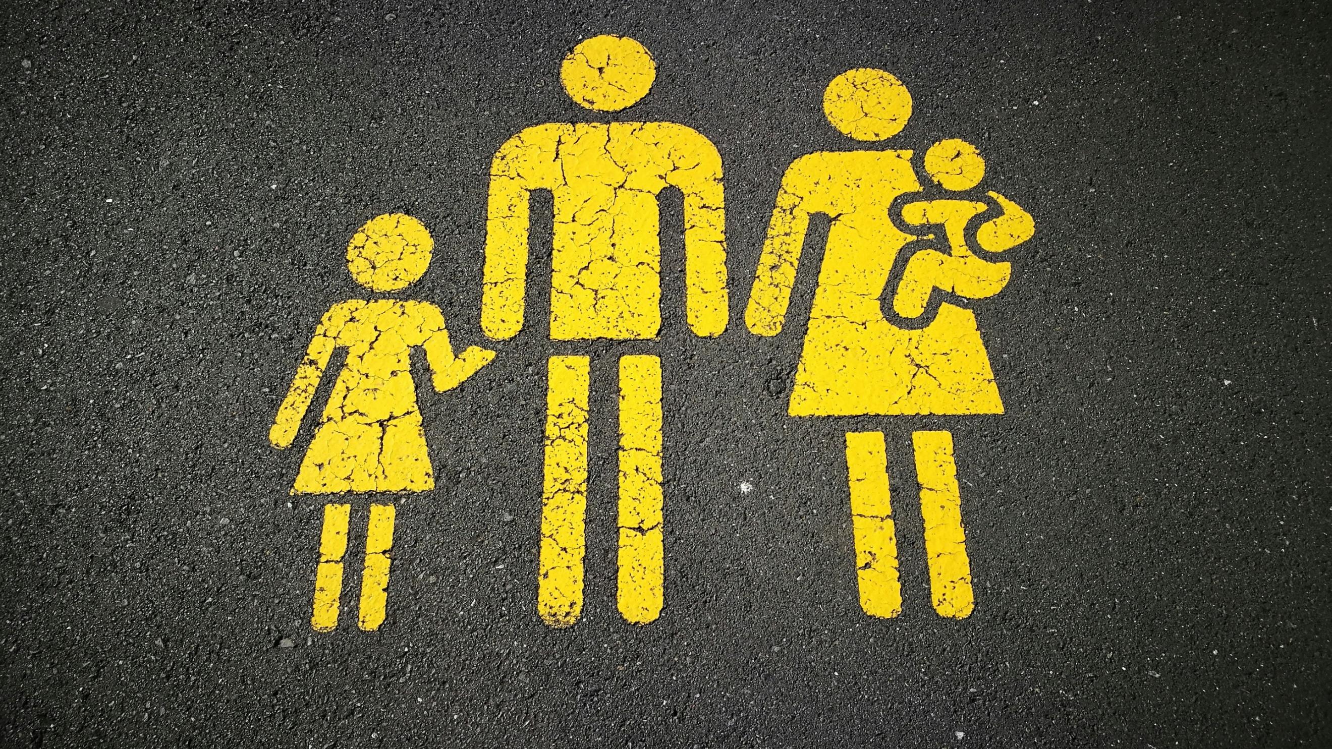 Auf einer Straße sind gelbe Strichmännchen aufgezeichnet. Es ist eine Familie von denen der Mann ein Kind an der Hand hält und die Frau ein Kind auf dem Arm (c) Sandy Millar / unsplash Auf einer Straße sind gelbe Strichmännchen aufgezeichnet. Es ist eine Familie von denen der Mann ein Kind an der Hand hält und die Frau ein Kind auf dem Arm
