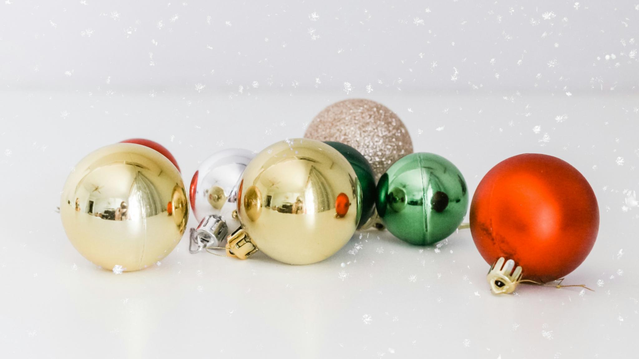 Auf einer weißen Oberfläche liegen Christbaumkugeln in gold, silber, rot und grün. Man sieht Schnee rieseln