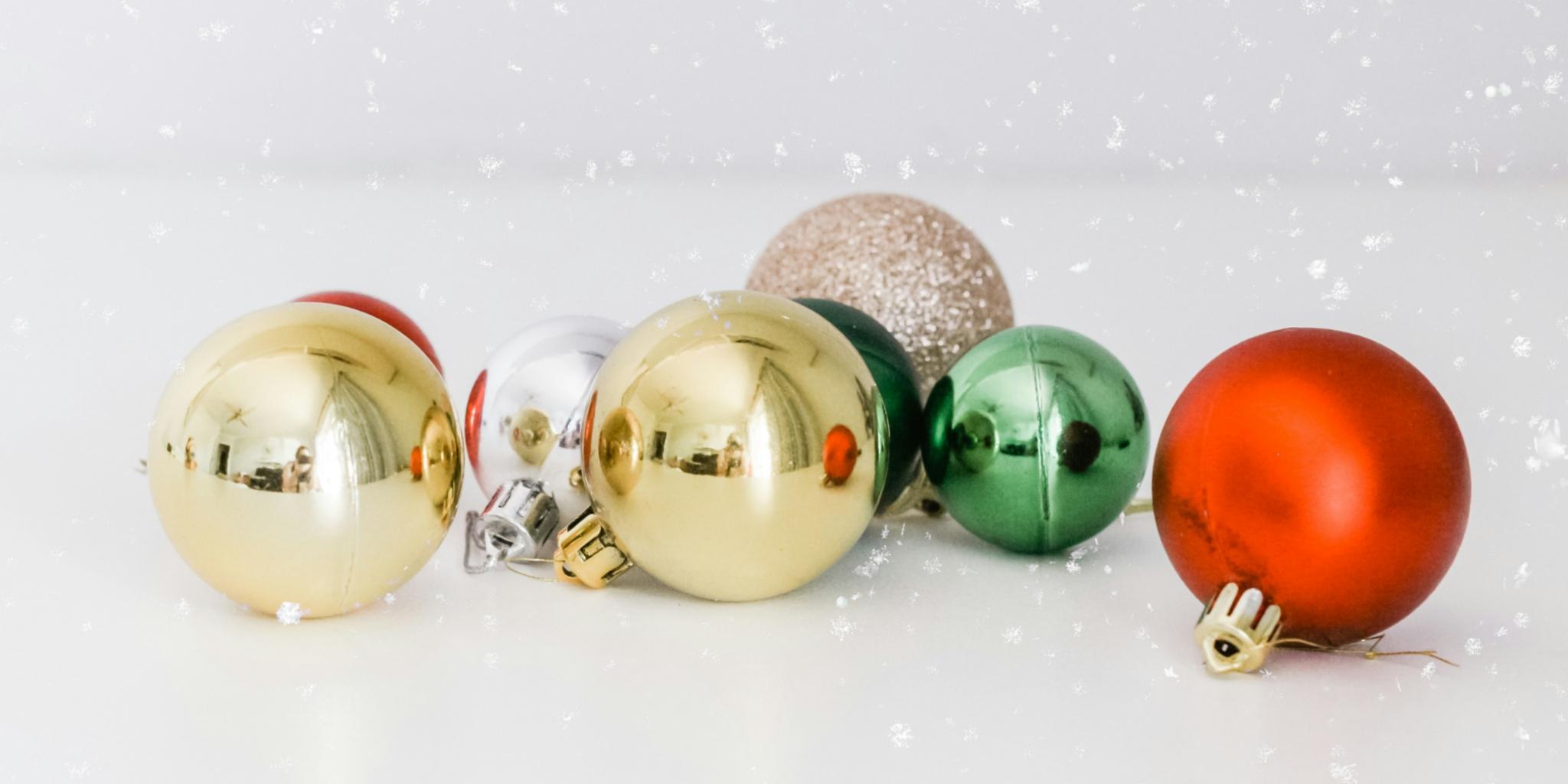 Auf einer weißen Oberfläche liegen Christbaumkugeln in gold, silber, rot und grün. Man sieht Schnee rieseln