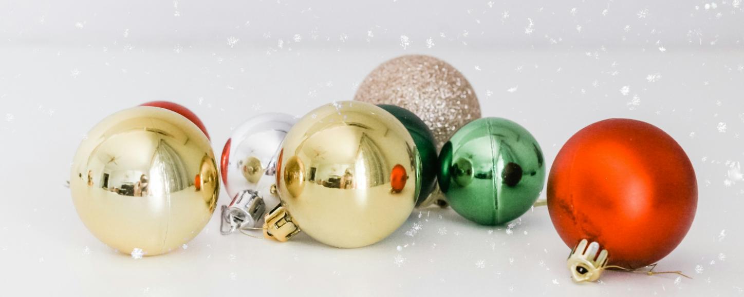 Auf einer weißen Oberfläche liegen Christbaumkugeln in gold, silber, rot und grün. Man sieht Schnee rieseln
