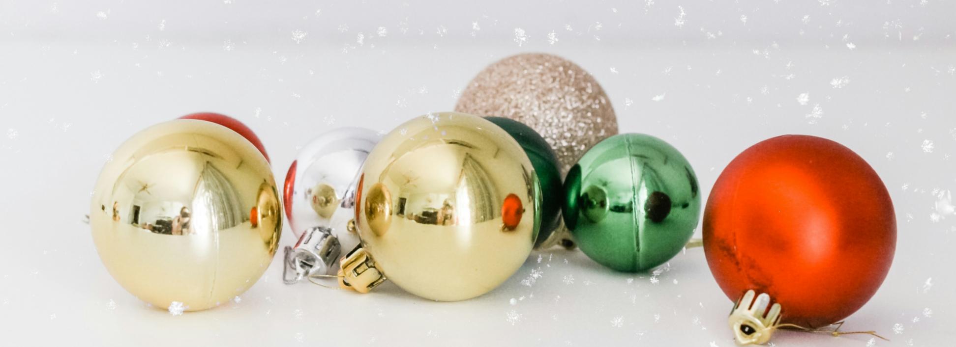 Auf einer weißen Oberfläche liegen Christbaumkugeln in gold, silber, rot und grün. Man sieht Schnee rieseln
