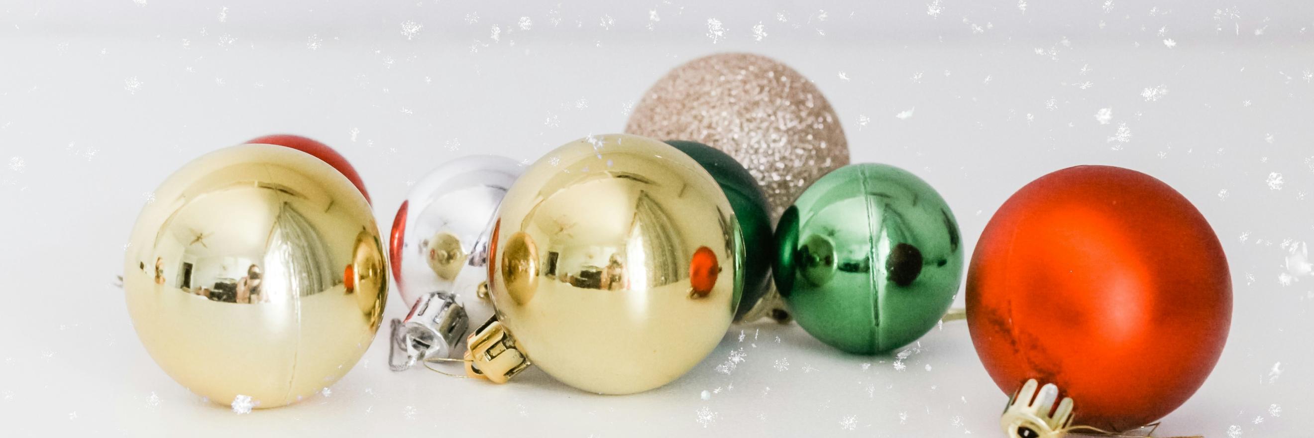 Auf einer weißen Oberfläche liegen Christbaumkugeln in gold, silber, rot und grün. Man sieht Schnee rieseln
