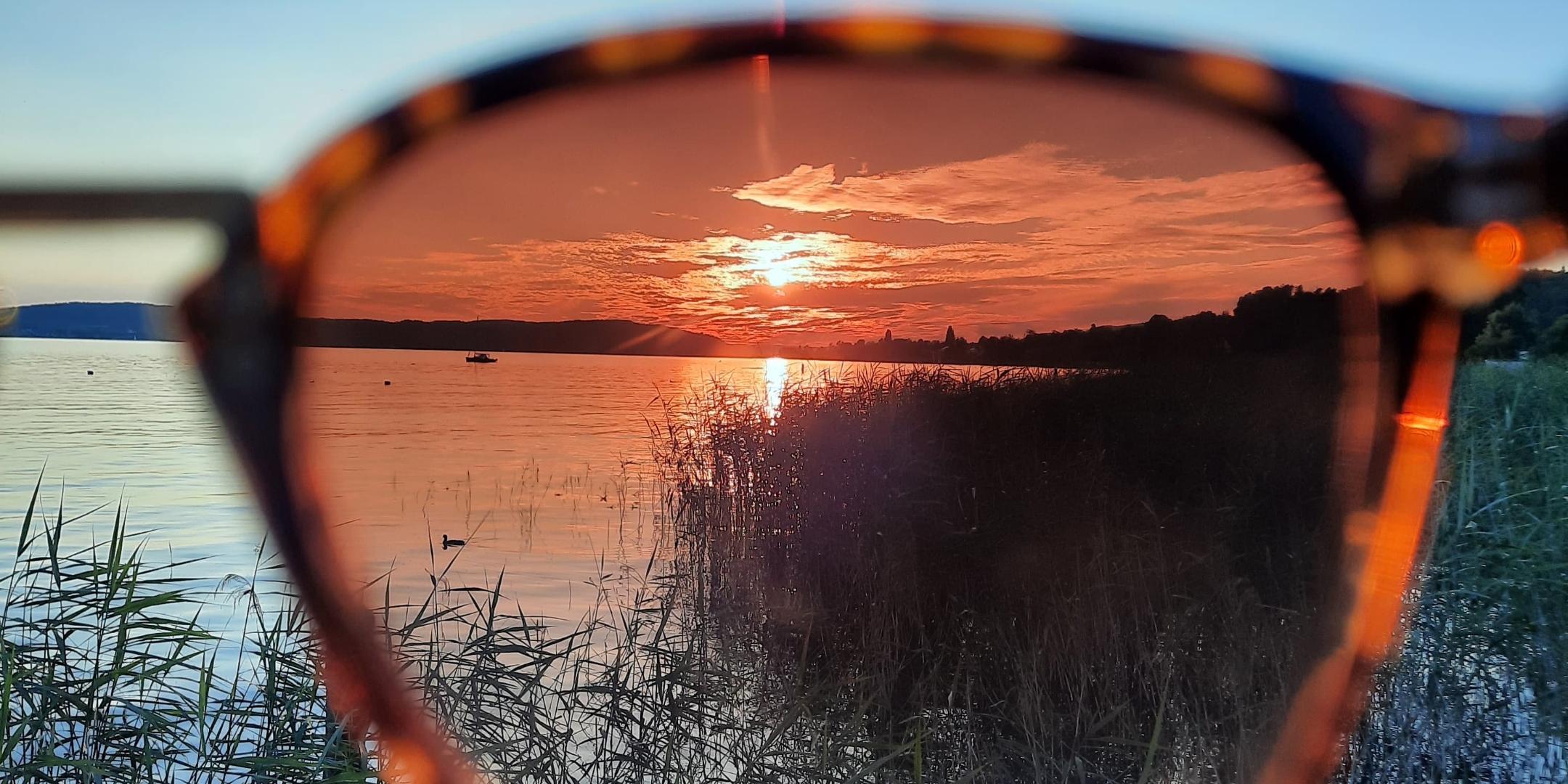 Man sieht einen See mit strahlend blauem Himmel und untergehender Sonne. Ein Teil des Bildes ist durch eine Sonnenbrille aufgenommen und wirkt dadurch dunkler und rötlich.