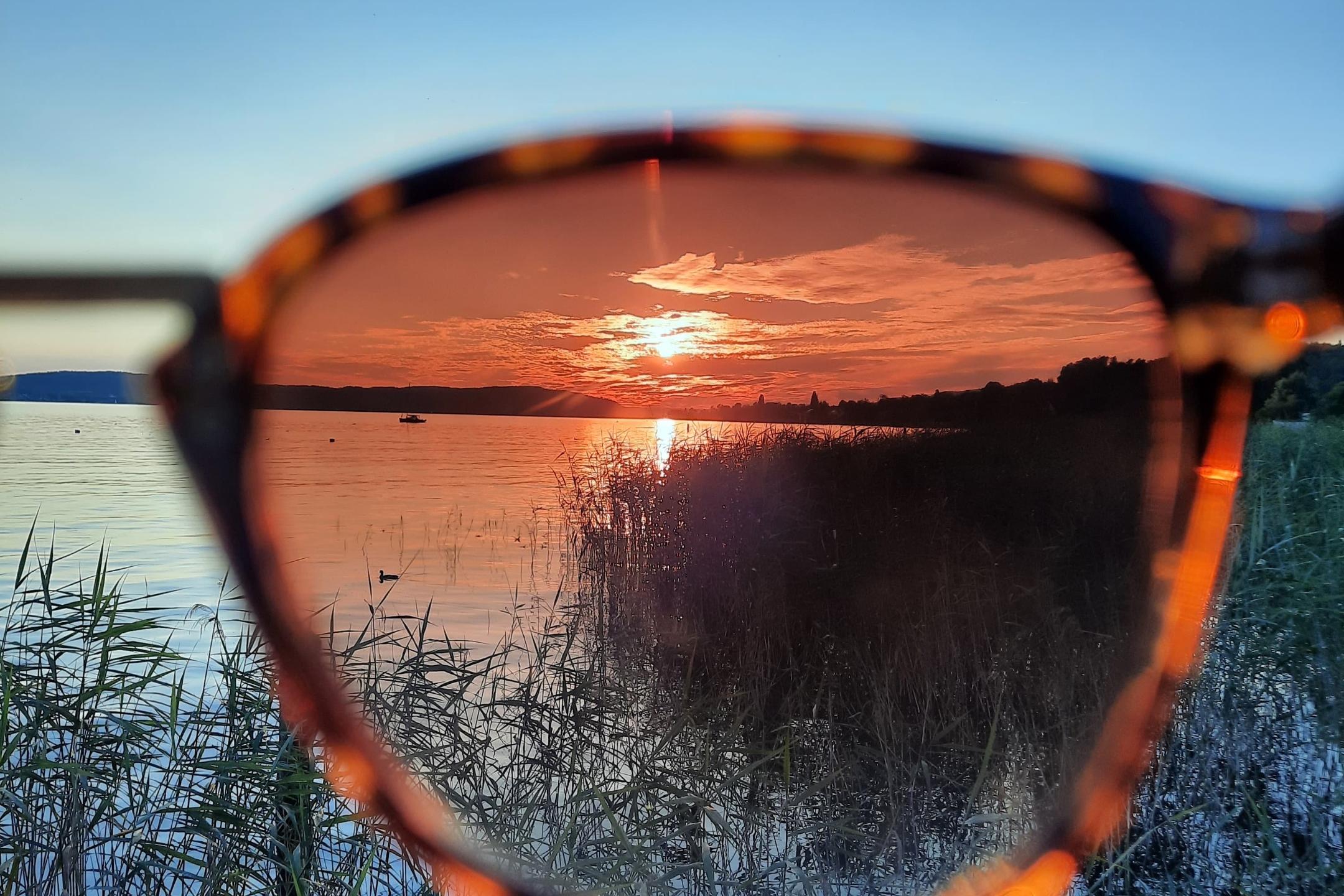 Man sieht einen See mit strahlend blauem Himmel und untergehender Sonne. Ein Teil des Bildes ist durch eine Sonnenbrille aufgenommen und wirkt dadurch dunkler und rötlich.