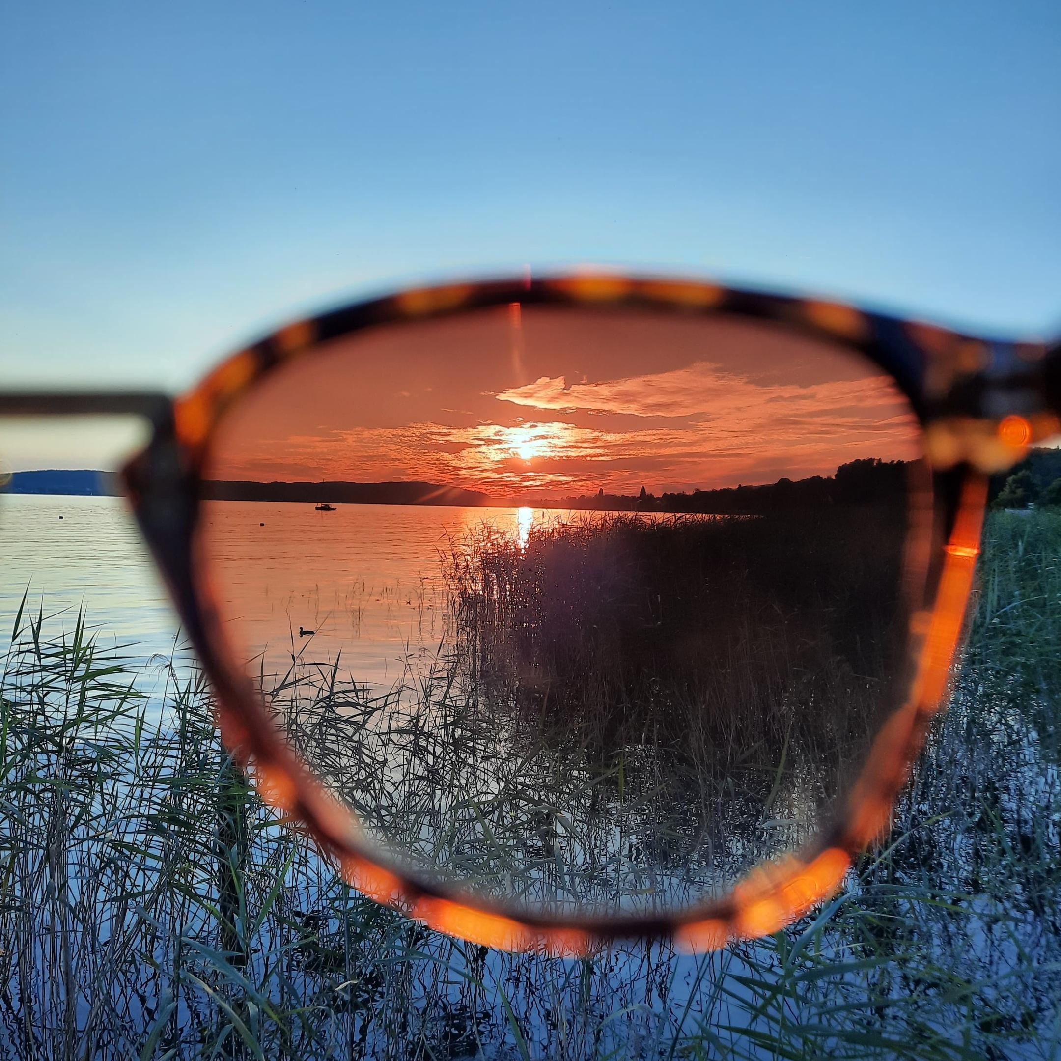 Man sieht einen See mit strahlend blauem Himmel und untergehender Sonne. Ein Teil des Bildes ist durch eine Sonnenbrille aufgenommen und wirkt dadurch dunkler und rötlich.