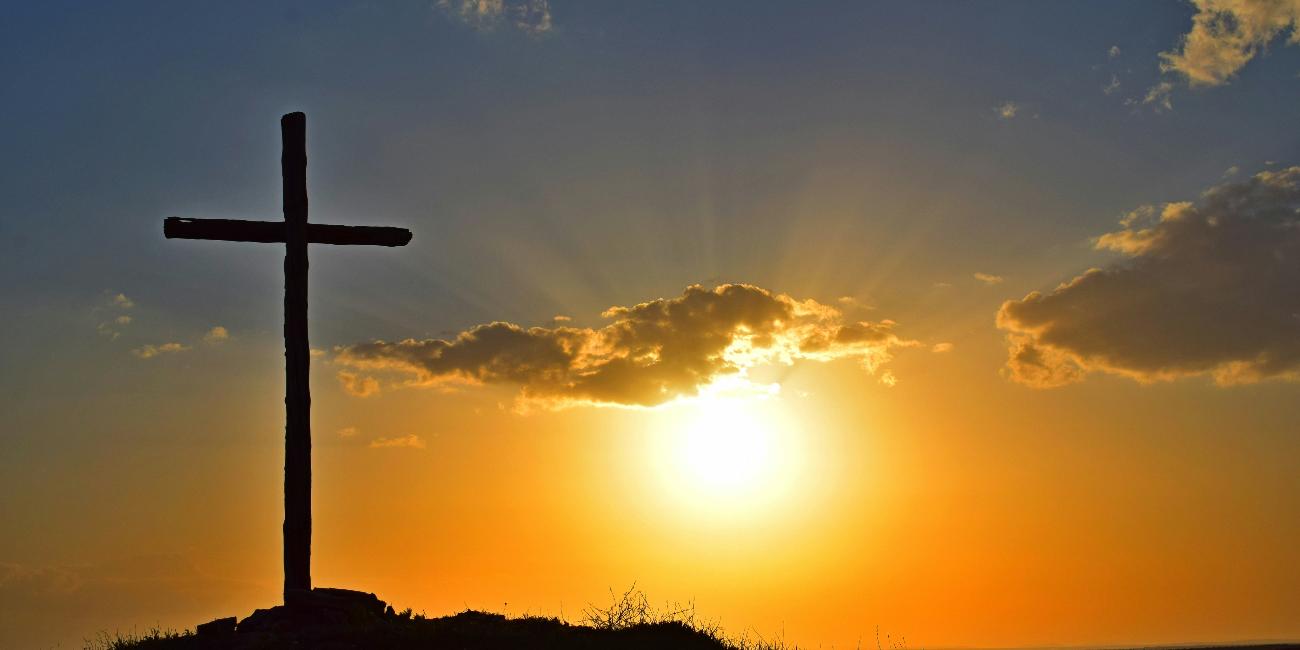 Blick in einen Sonnenuntergang. Auf einem kleinen Hügel steht in Kreuz, es ist nur als Silhouette zu sehen