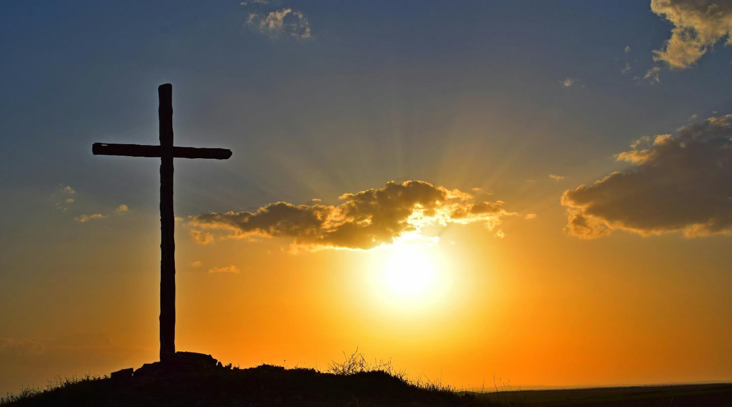 Blick in einen Sonnenuntergang. Auf einem kleinen Hügel steht in Kreuz, es ist nur als Silhouette zu sehen