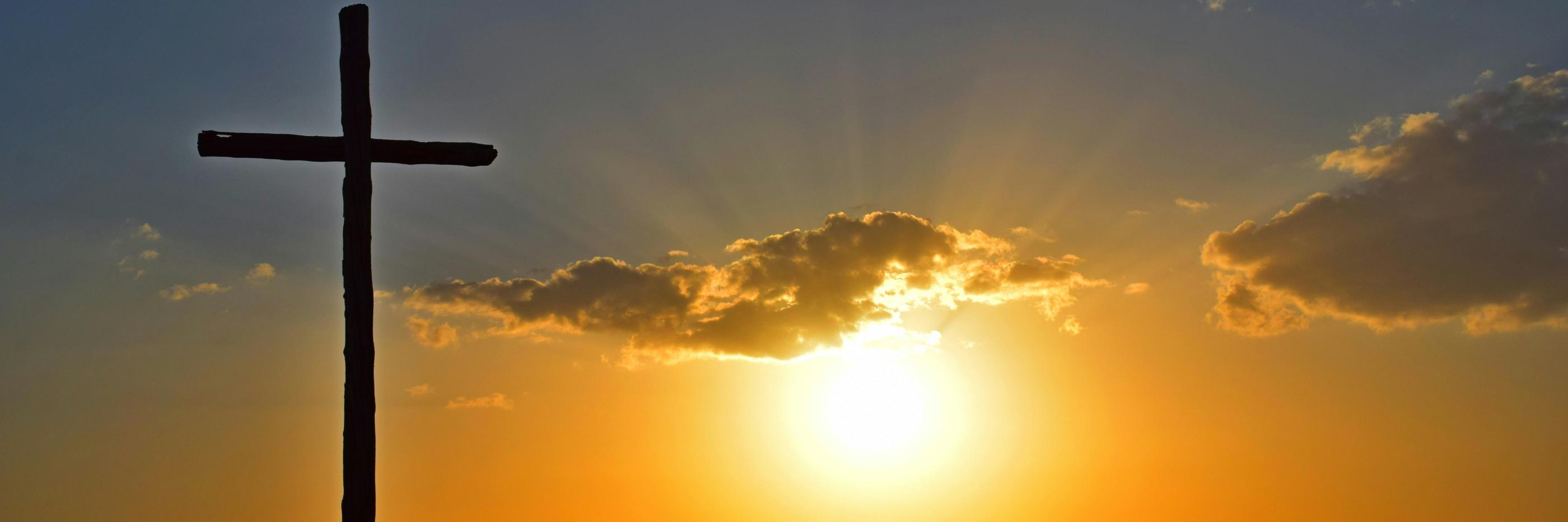 Blick in einen Sonnenuntergang. Auf einem kleinen Hügel steht in Kreuz, es ist nur als Silhouette zu sehen