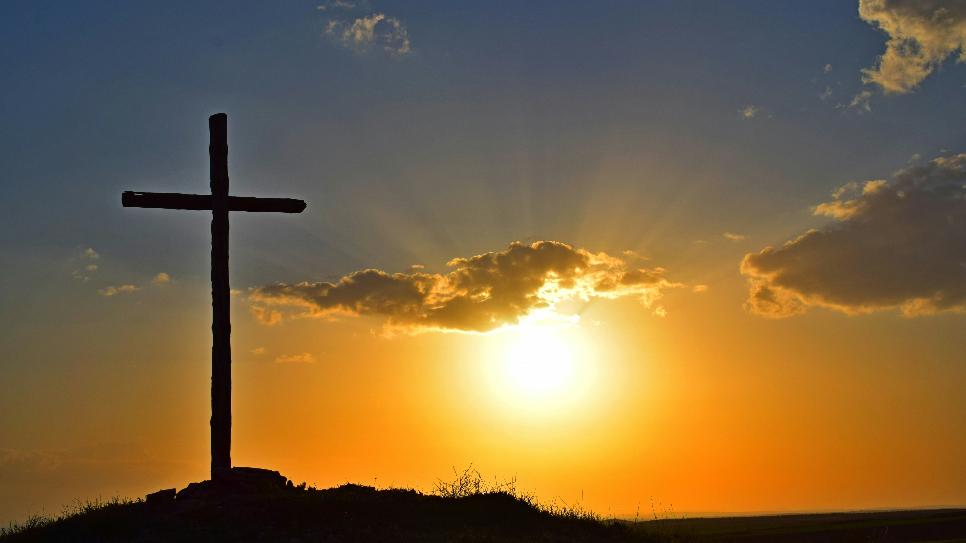 Blick in einen Sonnenuntergang. Auf einem kleinen Hügel steht in Kreuz, es ist nur als Silhouette zu sehen