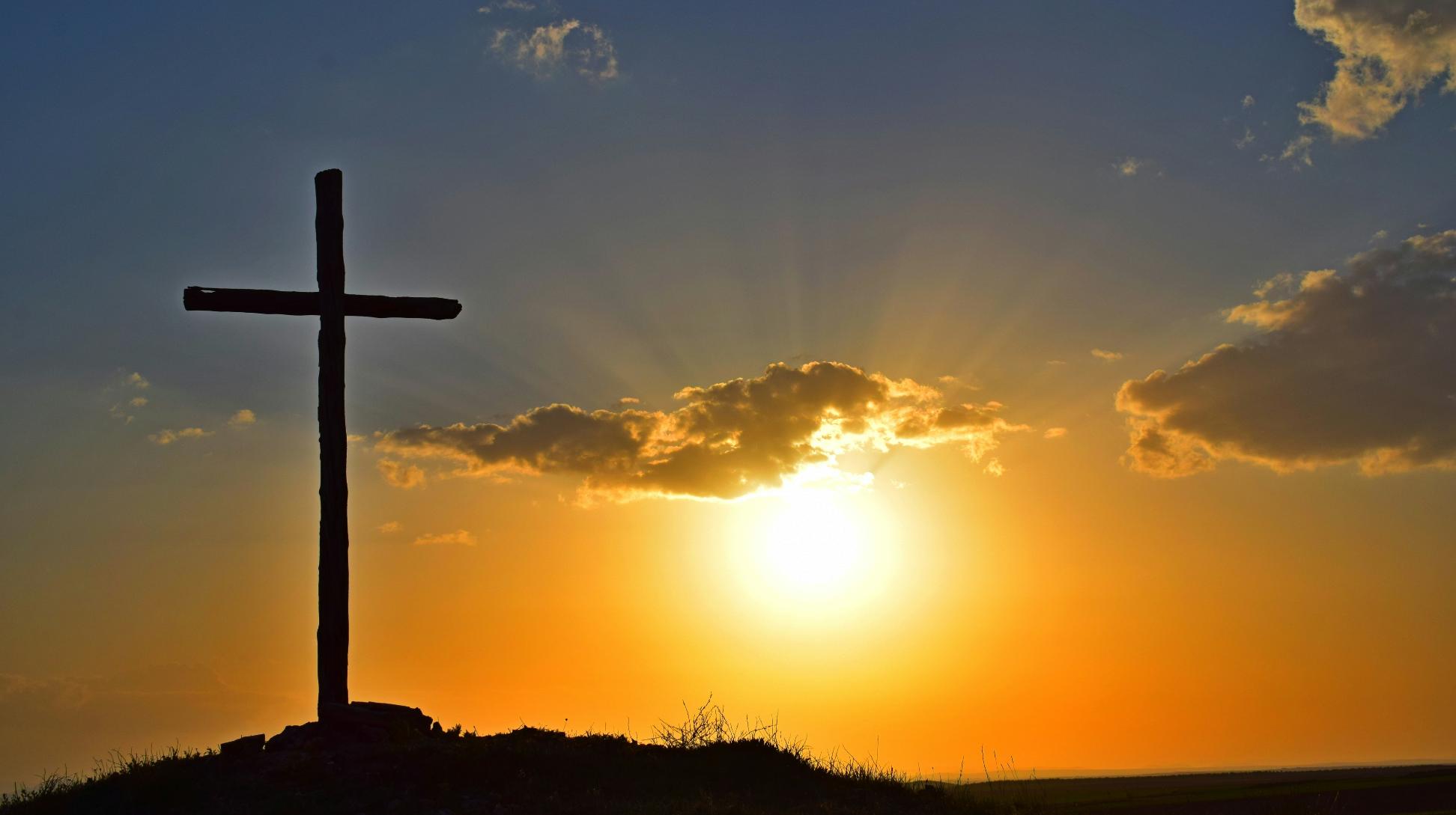 Blick in einen Sonnenuntergang. Auf einem kleinen Hügel steht in Kreuz, es ist nur als Silhouette zu sehen