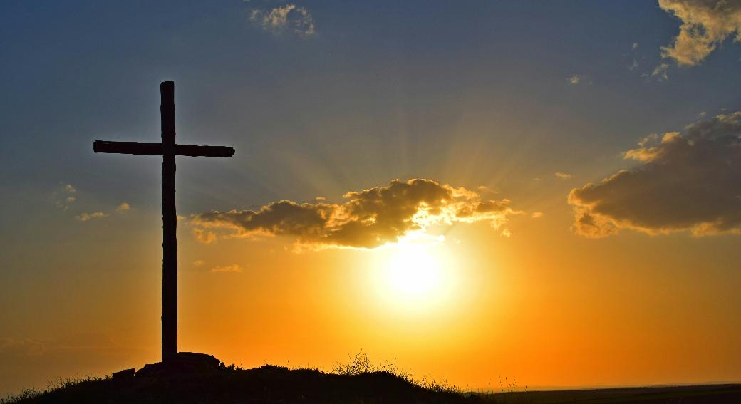 Blick in einen Sonnenuntergang. Auf einem kleinen Hügel steht in Kreuz, es ist nur als Silhouette zu sehen