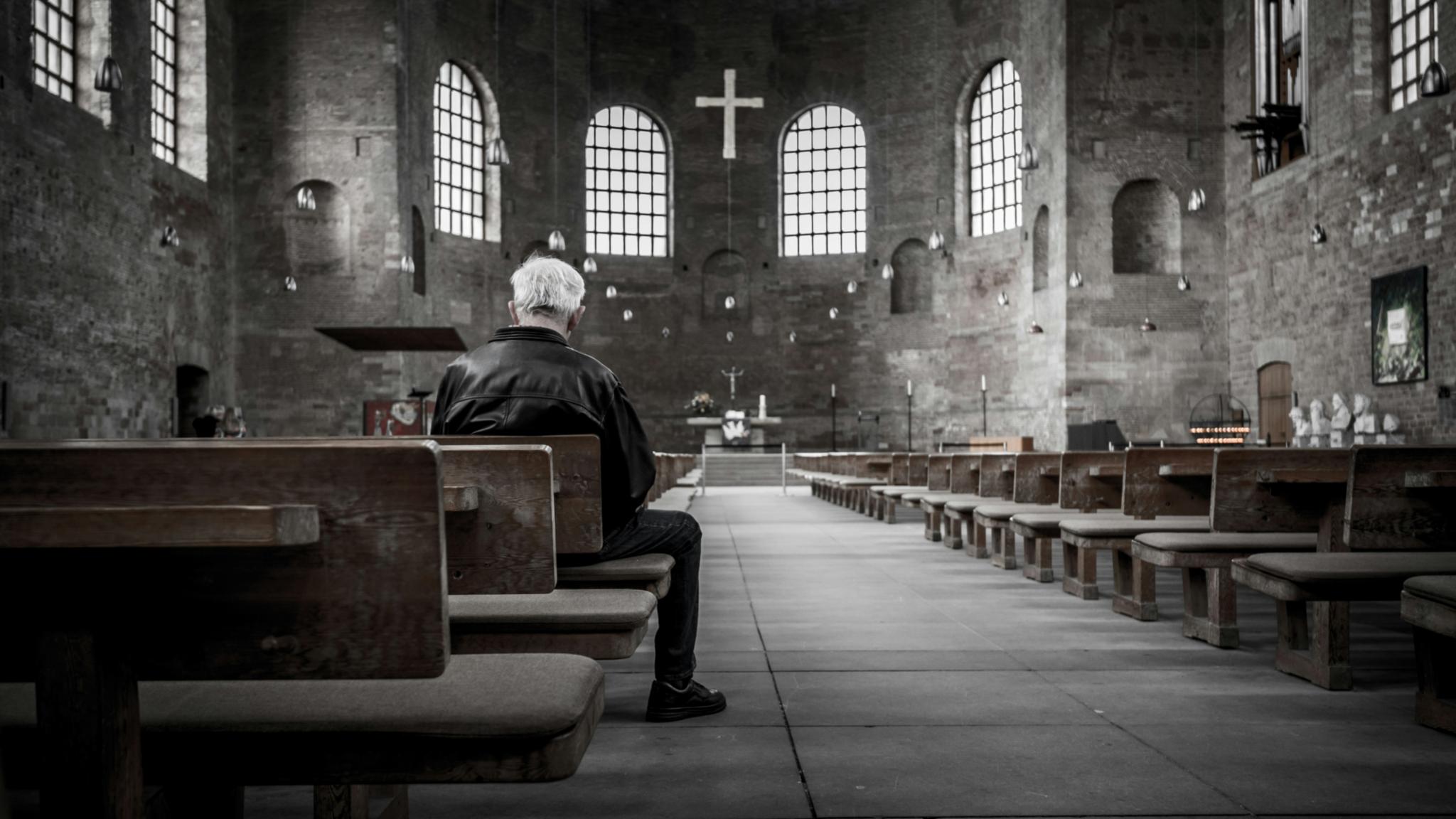 Ein Mann sitzt in einer Kircher auf einer Kirchenbank. Der Mann ist nur von hinten zu sehen. Das Bild ist schwarz weiß