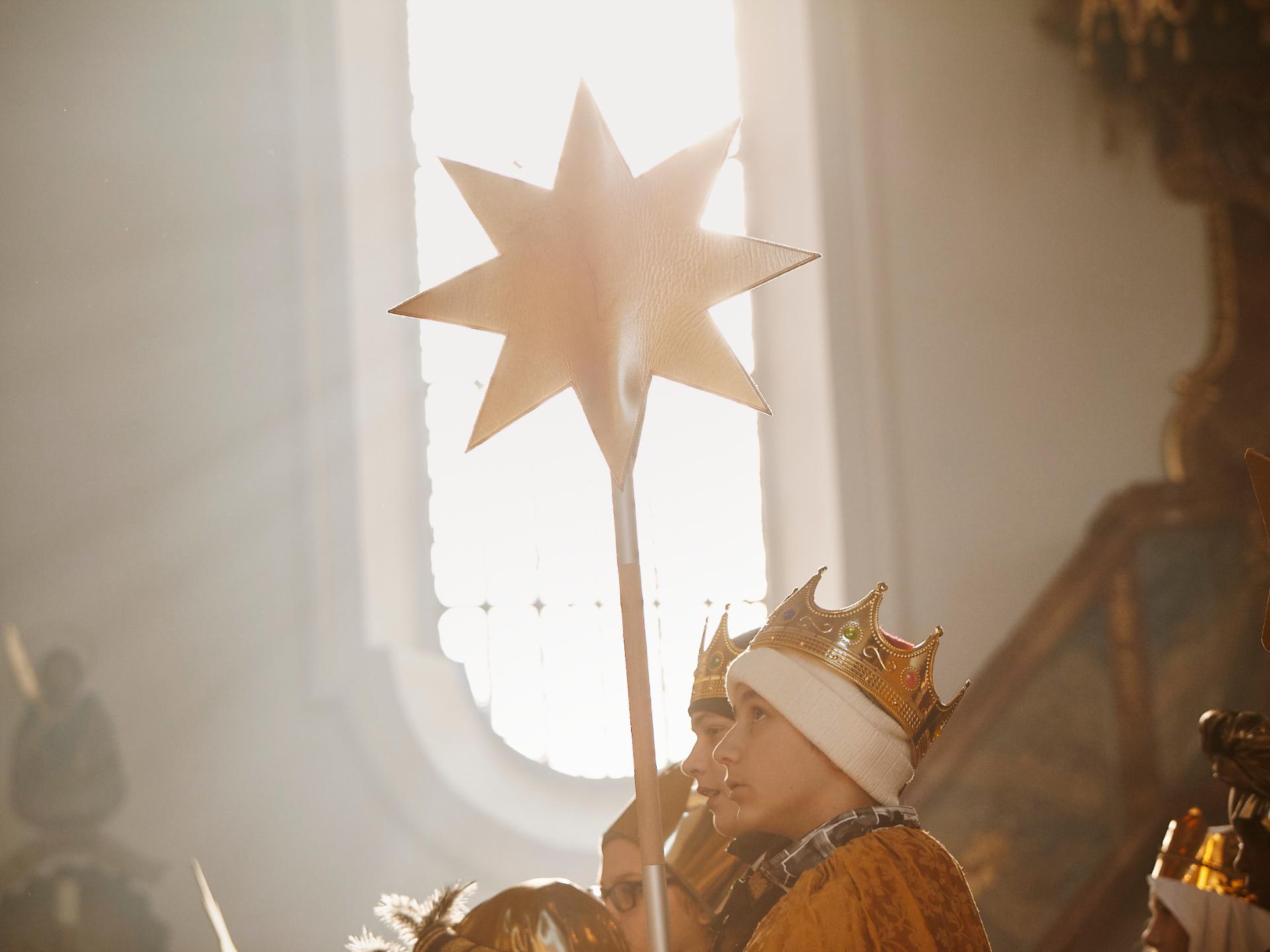 Blick auf ein weißes Kirchenfenster. Davor stehen Sternsinger, die einen Stern vor das Fenster halten