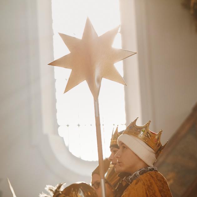 Blick auf ein weißes Kirchenfenster. Davor stehen Sternsinger, die einen Stern vor das Fenster halten