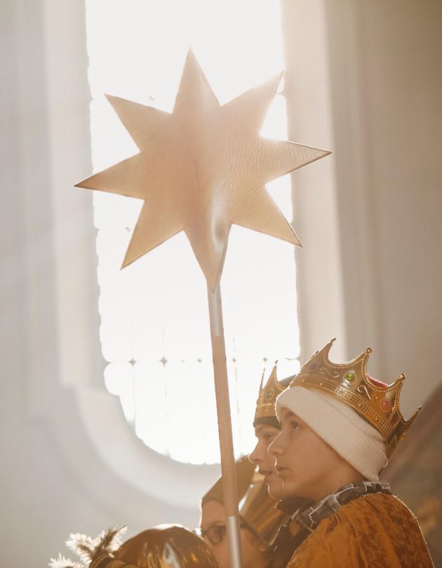 Blick auf ein weißes Kirchenfenster. Davor stehen Sternsinger, die einen Stern vor das Fenster halten