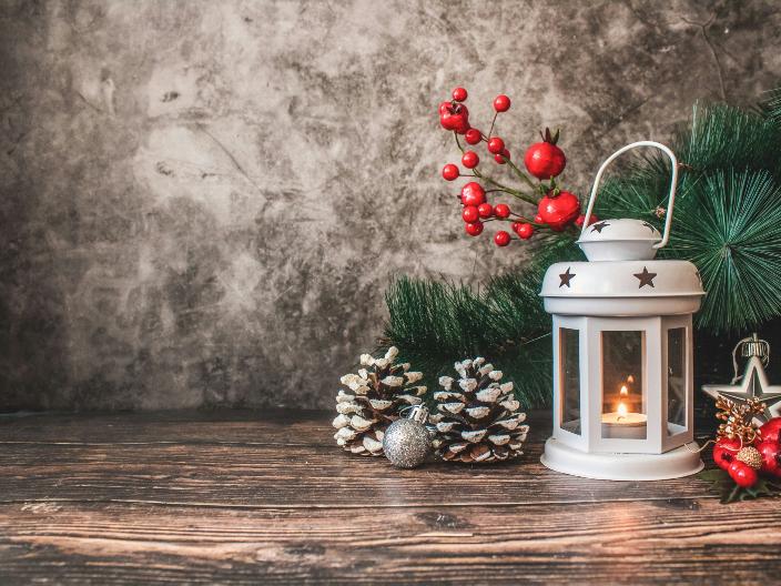 Auf einem Holztisch steht eine weiße weihnachtliche Laterne mit einer Kerze. Um die Laterne wurde mit Tannenzweigen, roten Beeren und Tannenzapfen dekoriert