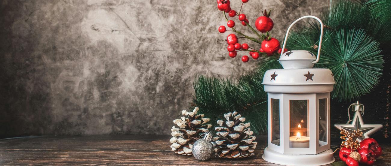 Auf einem Holztisch steht eine weiße weihnachtliche Laterne mit einer Kerze. Um die Laterne wurde mit Tannenzweigen, roten Beeren und Tannenzapfen dekoriert