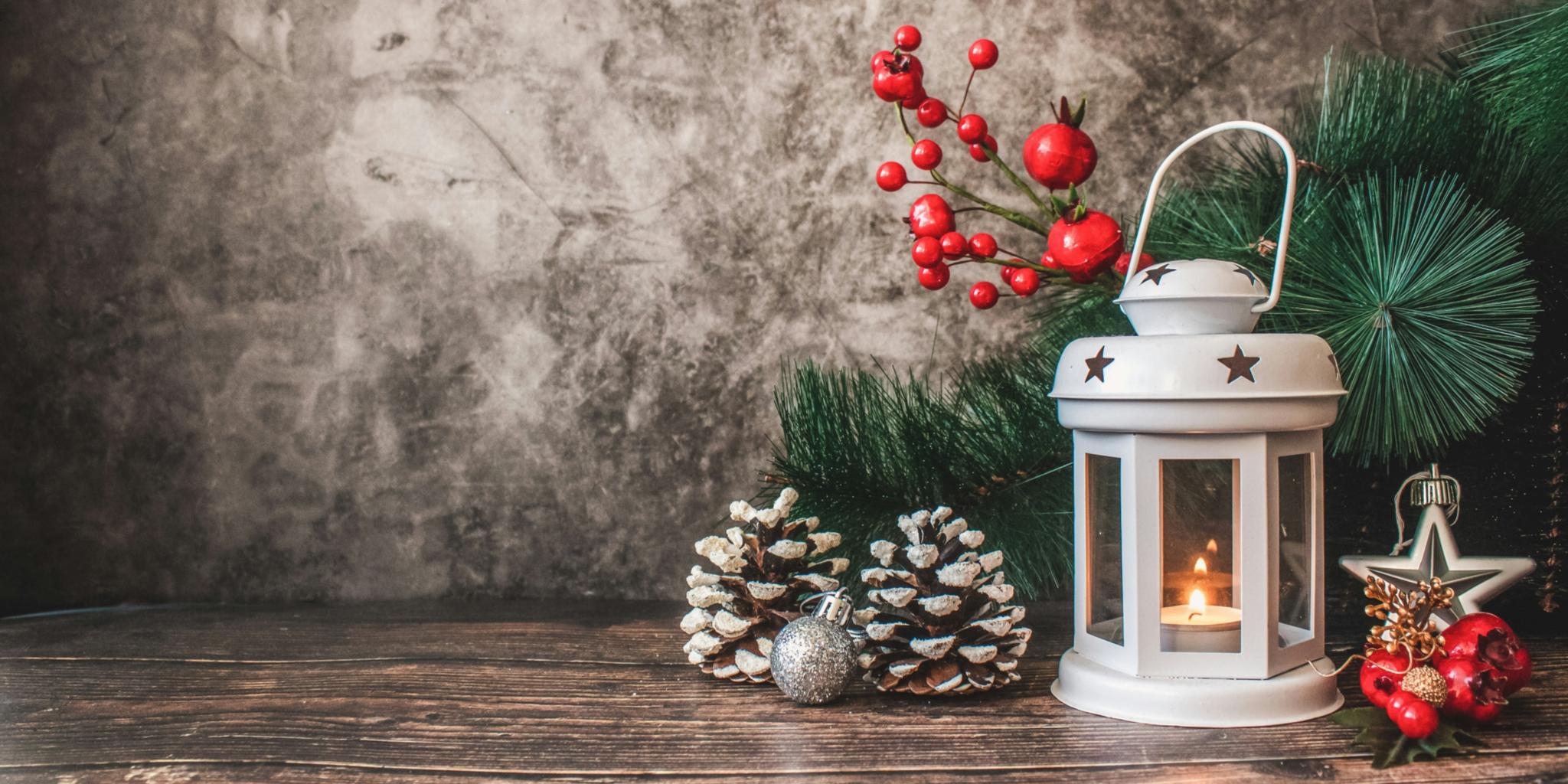 Auf einem Holztisch steht eine weiße weihnachtliche Laterne mit einer Kerze. Um die Laterne wurde mit Tannenzweigen, roten Beeren und Tannenzapfen dekoriert