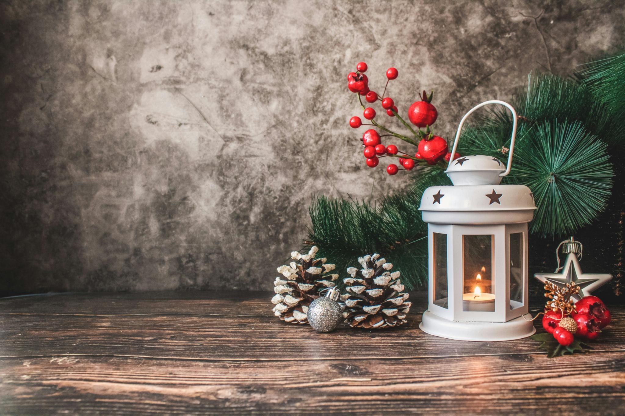 Auf einem Holztisch steht eine weiße weihnachtliche Laterne mit einer Kerze. Um die Laterne wurde mit Tannenzweigen, roten Beeren und Tannenzapfen dekoriert