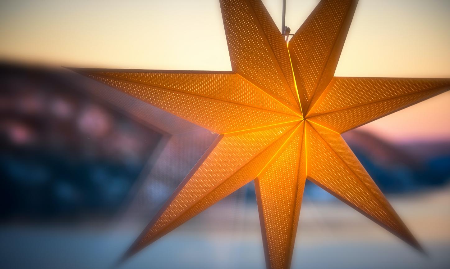 Man sieht einen leuchtenden Stern an einer Fensterscheibe. Im Hintergrund ein Sonnenauf- oder untergang