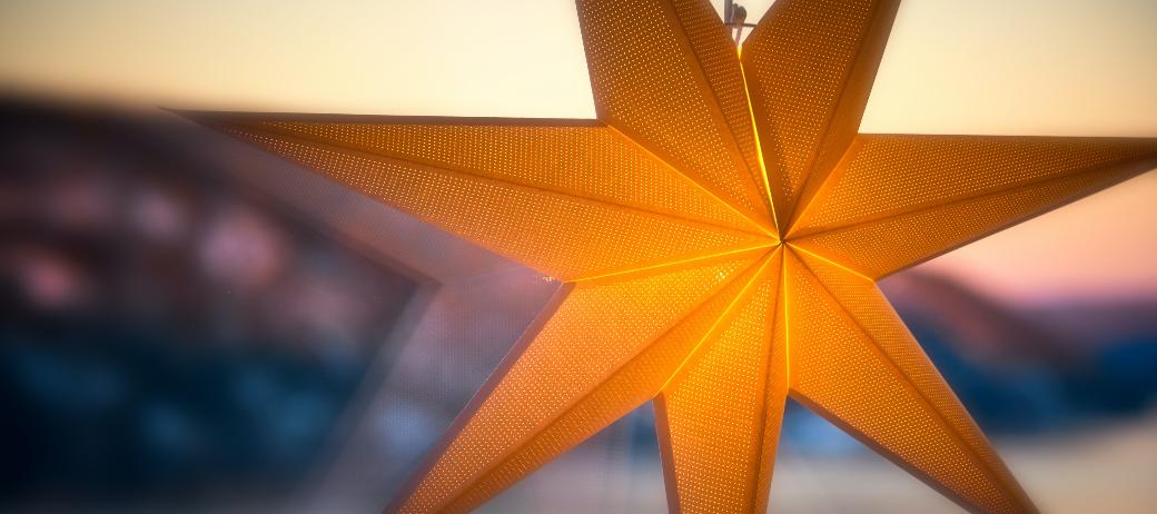 Man sieht einen leuchtenden Stern an einer Fensterscheibe. Im Hintergrund ein Sonnenauf- oder untergang