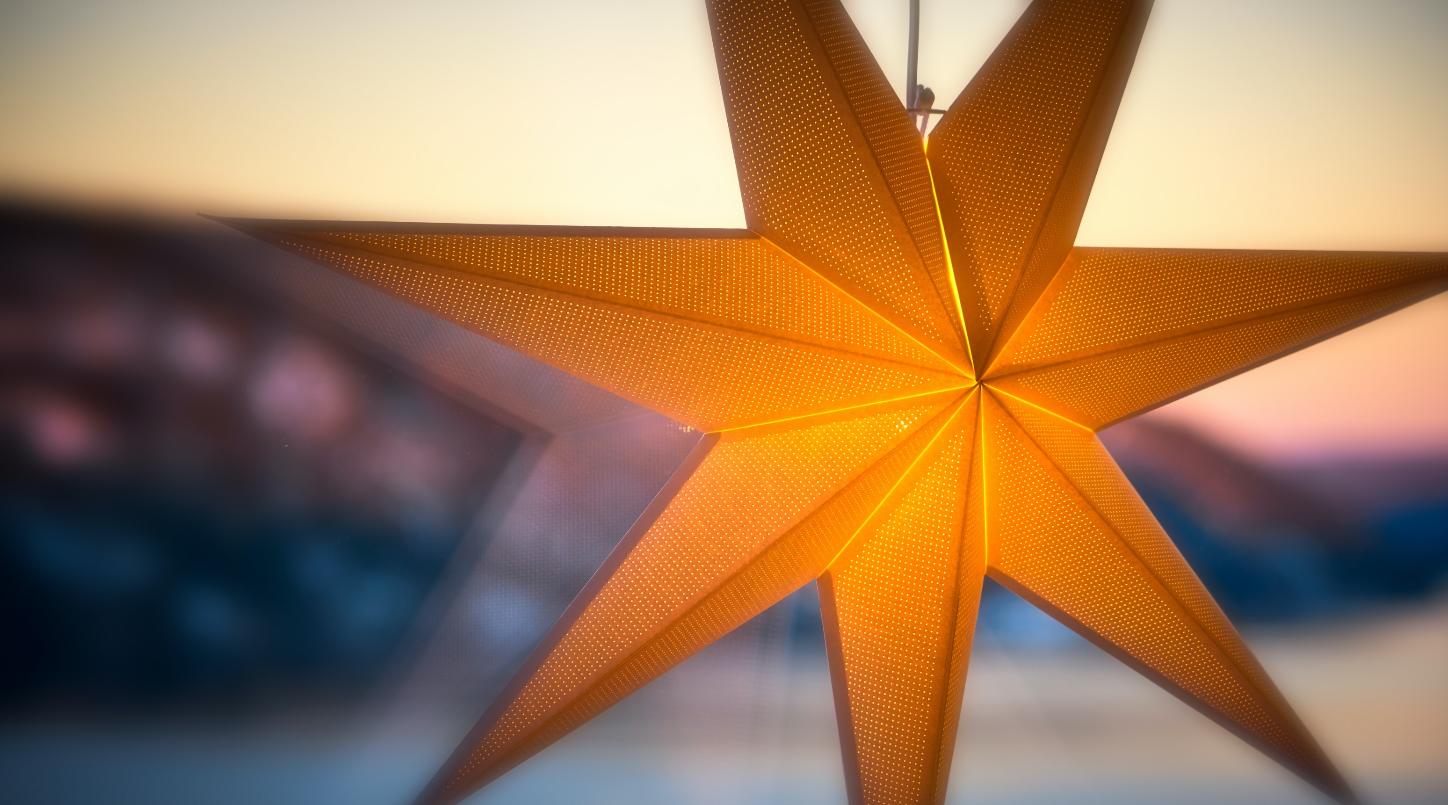 Man sieht einen leuchtenden Stern an einer Fensterscheibe. Im Hintergrund ein Sonnenauf- oder untergang