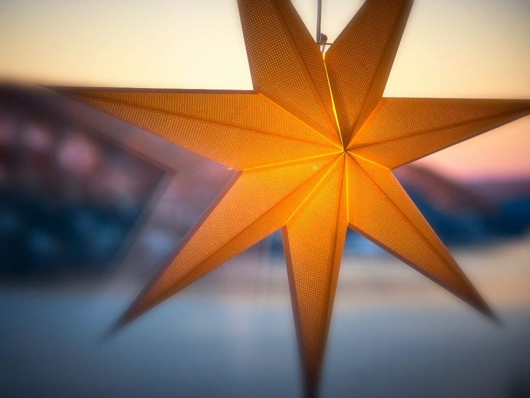 Man sieht einen leuchtenden Stern an einer Fensterscheibe. Im Hintergrund ein Sonnenauf- oder untergang