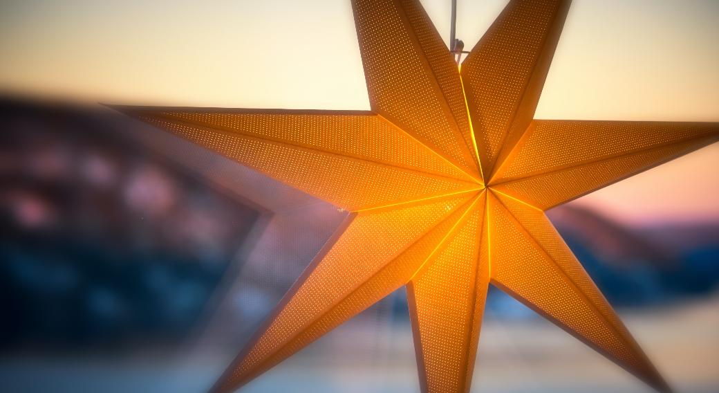 Man sieht einen leuchtenden Stern an einer Fensterscheibe. Im Hintergrund ein Sonnenauf- oder untergang