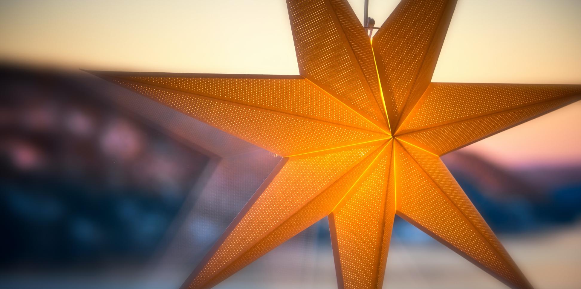 Man sieht einen leuchtenden Stern an einer Fensterscheibe. Im Hintergrund ein Sonnenauf- oder untergang