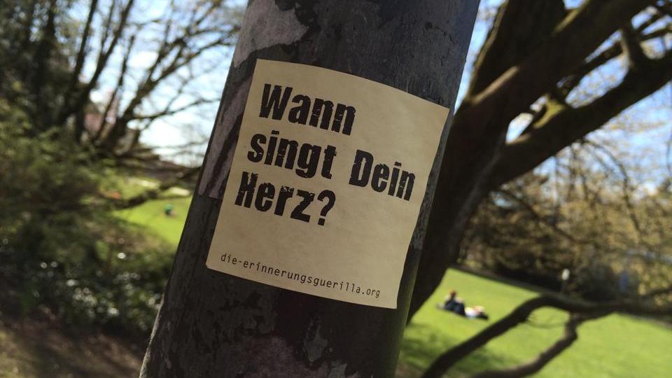 Ein Aufkleber mit dem Text 'Wann singt Dein Herz?' klebt auf einem Laternenpfahl