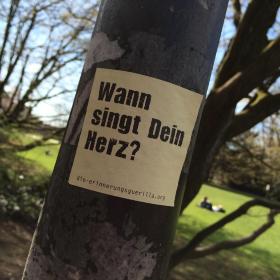 Ein Aufkleber mit dem Text 'Wann singt Dein Herz?' klebt auf einem Laternenpfahl