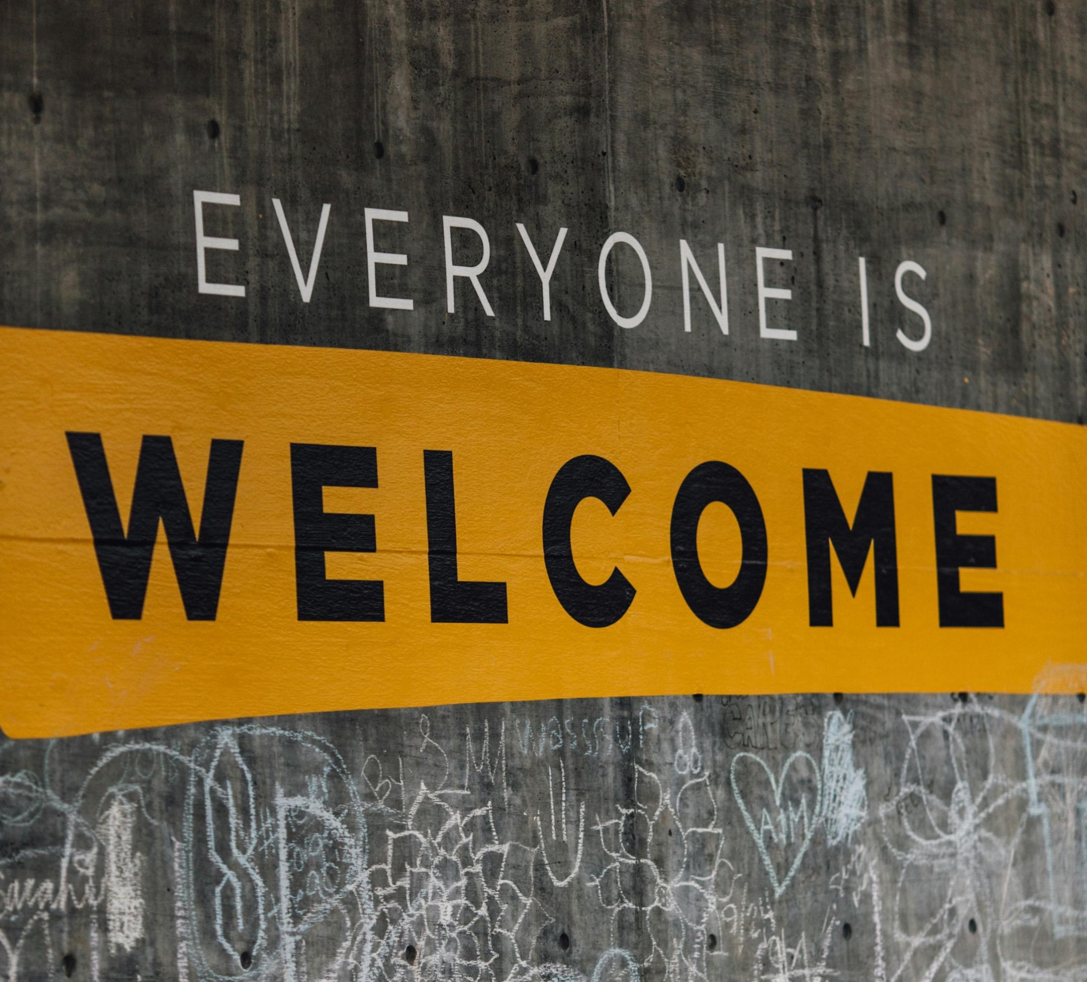 Willkommen! (c) Katie Moum auf Unsplash Auf einer Wand steht der Text 'Everyone is welcome'. Darunter sind viele kleine Kreidezeichnungen