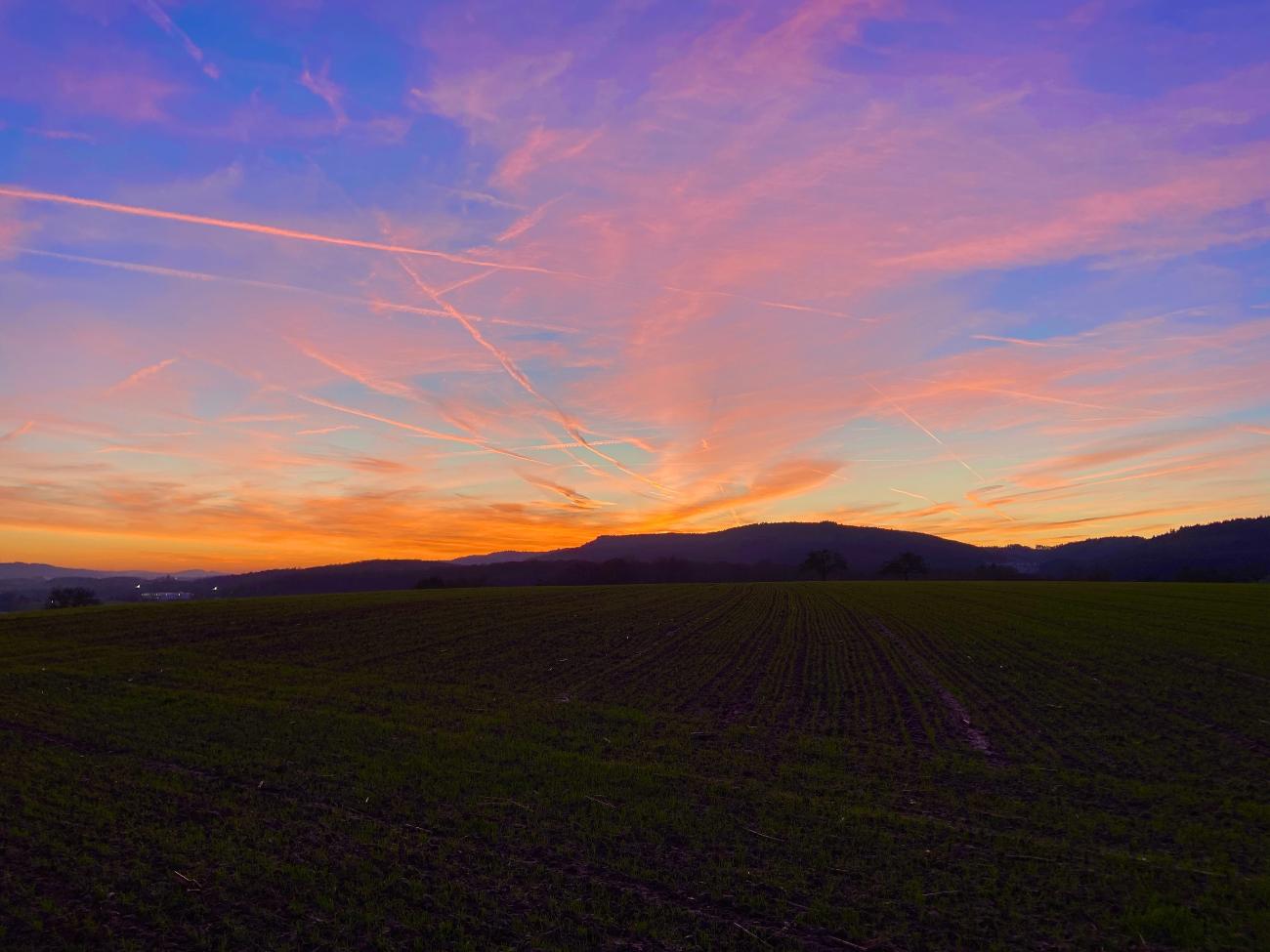 Blick auf einen dramatischen Sonnenuntergang am Horizont. Am Himmel werden Kondensstreifen orange gefärbt