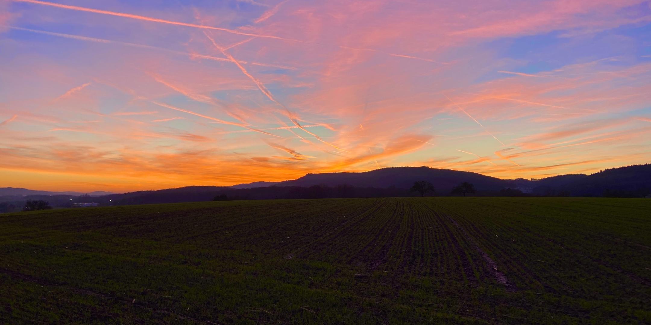Blick auf einen dramatischen Sonnenuntergang am Horizont. Am Himmel werden Kondensstreifen orange gefärbt