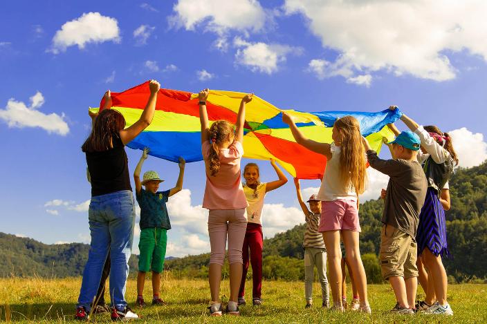 Eine Gruppe Kinder hält ein buntes 'Schwungtuch' in die Luft. Sie stehen auf einer Wiese. Der Himmel ist blau-weiß bewölkt