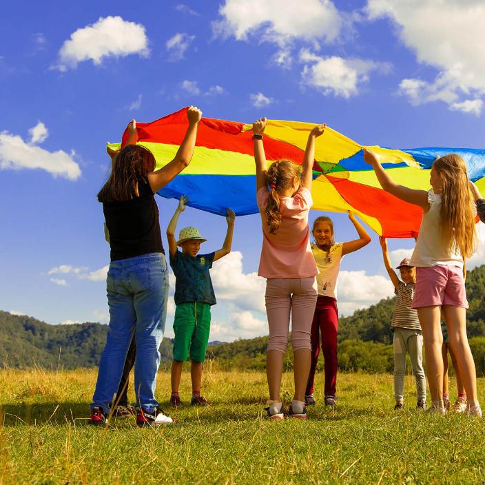 Eine Gruppe Kinder hält ein buntes 'Schwungtuch' in die Luft. Sie stehen auf einer Wiese. Der Himmel ist blau-weiß bewölkt